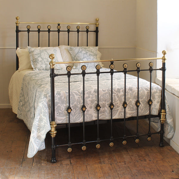 A black metal bed frame with ornate gold-toned accents and brass-finished posts, shown on a wooden floor in a bedroom setting with patterned bedding.