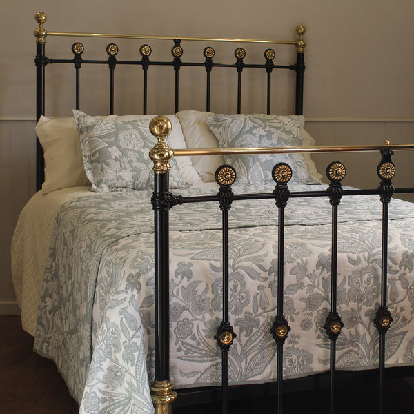 A classic black metal bed frame with ornate gold-toned accents and brass-finished posts, shown made up with a light gray and white damask patterned duvet and matching pillows in a bedroom setting.