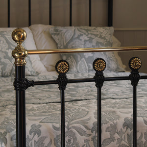 Close-up of a classic black metal bed frame with ornate gold-toned accents, shown with patterned bedding and pillows in a bedroom setting.