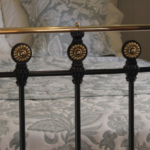 Close-up of a bed frame with ornate black metal posts topped with gold-toned circular finials, shown against patterned bedding and pillows.