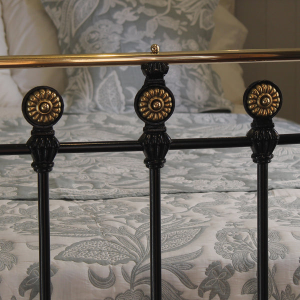 Close-up of a bed frame with ornate black metal posts topped with gold-toned circular finials, shown against patterned bedding and pillows.