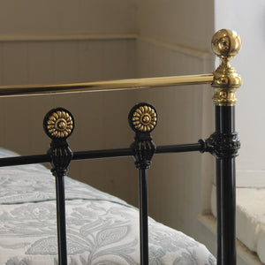 Close-up of a black metal bed frame with ornate gold-finished decorative accents and a matching gold horizontal rail, shown in a bedroom setting with patterned bedding.