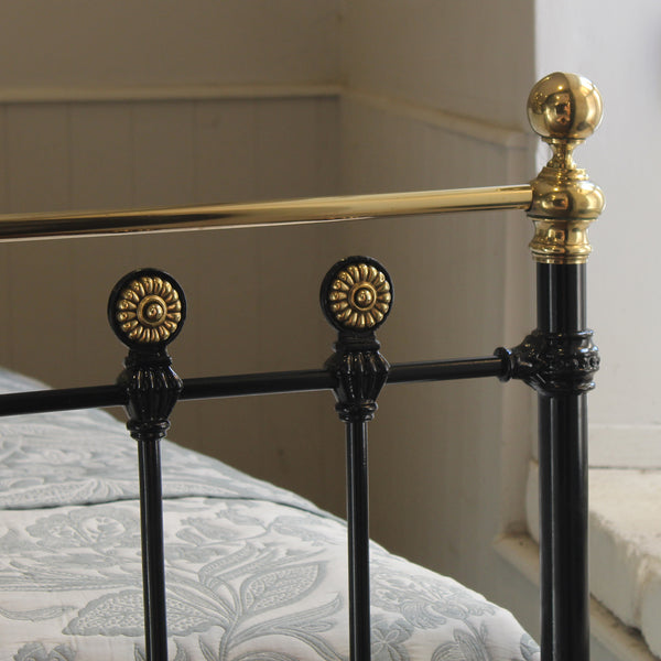 Close-up of a black metal bed frame with ornate gold-finished decorative accents and a matching gold horizontal rail, shown in a bedroom setting with patterned bedding.