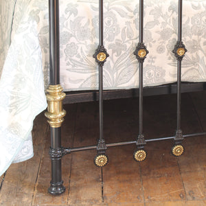Close-up of a black metal bed frame with ornate gold-toned decorative accents and a floral-patterned cream bedspread, set on a rustic wooden floor.