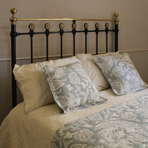 A styled bed with a black metal headboard featuring gold accents, dressed in cream bedding and two decorative light blue-gray pillows with a white floral damask pattern.