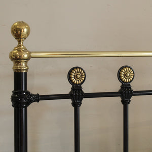 Close-up of an ornate metal bed frame showing a black painted iron body with polished gold-finished decorative accents, including a spherical finial, a horizontal rail, and circular rosette details.