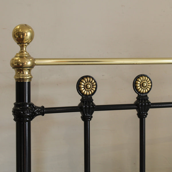Close-up of an ornate metal bed frame showing a black painted iron body with polished gold-finished decorative accents, including a spherical finial, a horizontal rail, and circular rosette details.