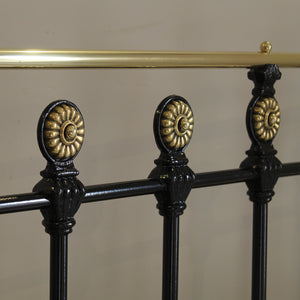 Close-up of a black metal bed with ornate gold-toned circular finials and a brass-colored top rail against a plain beige wall.