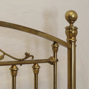 Close-up of a polished brass bed frame showing the headboard corner with a spherical finial, curved rail, and decorative scrollwork against a textured off-white wall.