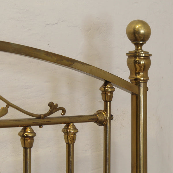 Close-up of a polished brass bed frame showing the headboard corner with a spherical finial, curved rail, and decorative scrollwork against a textured off-white wall.