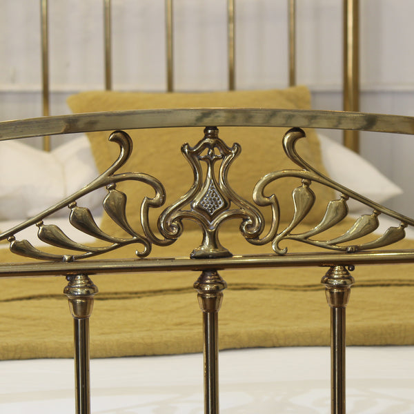 Close-up of an ornate brass bed frame headboard with decorative scrollwork and vertical bars, shown in a bedroom setting with a yellow upholstered headboard panel and white bedding behind it.