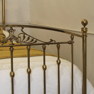 Close-up of a polished brass metal bed frame with decorative scrollwork on the headboard and vertical finials, shown with white bedding and a yellow pillow in a bedroom setting.