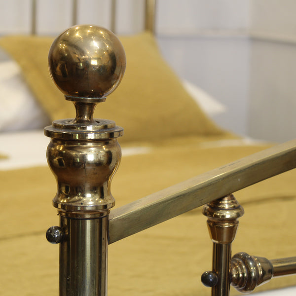 Close-up of a polished brass bed frame post with a spherical finial and matching metal rail, set against a softly blurred bed with yellow bedding.