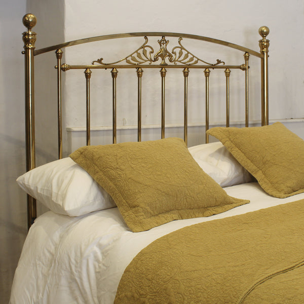 A brass metal bed frame with an ornate crest pattern on the headboard, styled with white bedding, white pillows, and two textured mustard yellow accent pillows.