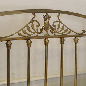 Close-up of a brass bed headboard with ornate scrollwork and decorative finials against a plain off-white wall.