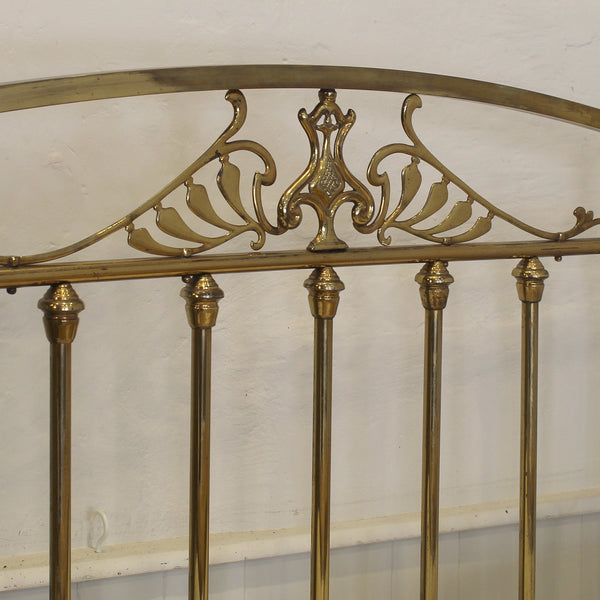 Close-up of a brass bed headboard with ornate scrollwork and decorative finials against a plain off-white wall.