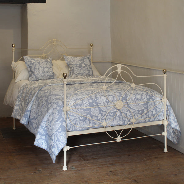 An antique white metal bed frame with ornate scrollwork, dressed in a blue and white floral patterned quilt and matching pillows, set in a room with a dark wood floor and neutral walls.