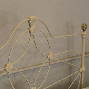 Close-up of an antique metal bed frame headboard in a cream finish, featuring decorative scrollwork and a gold-finished finial on the side post against a textured off-white wall.