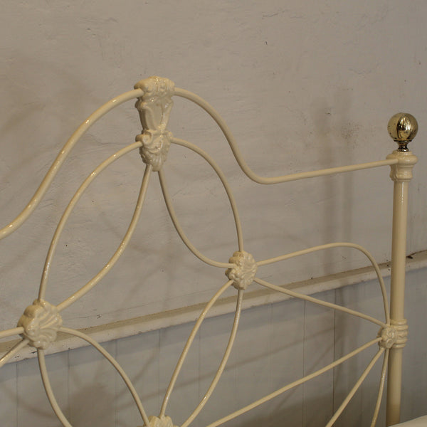 Close-up of an antique metal bed frame headboard in a cream finish, featuring decorative scrollwork and a gold-finished finial on the side post against a textured off-white wall.