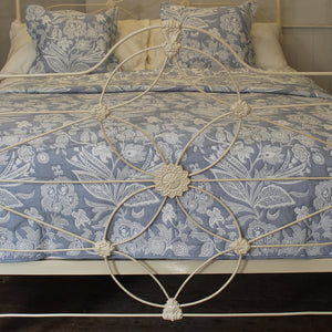 A close-up of a bed with a light blue and white floral patterned quilt and matching pillow shams, set against a white metal headboard with an ornate circular design.