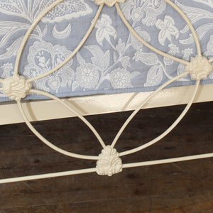 Close-up of a white metal bed frame with decorative scrollwork and a blue floral-patterned quilt on a dark wood floor.