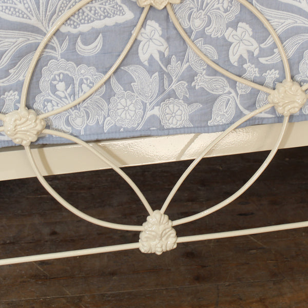 Close-up of a white metal bed frame with decorative scrollwork and a blue floral-patterned quilt on a dark wood floor.