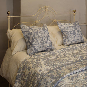 A neatly made bed with a cream metal headboard, featuring a blue and cream floral patterned comforter and two matching decorative pillows against plain cream pillows, set against a neutral wall background.
