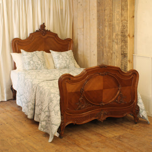 An ornate vintage-style wooden bed frame with a carved headboard and matching footboard, dressed in white and pale blue patterned bedding, set in a rustic room with wood-paneled walls and a wooden floor.
