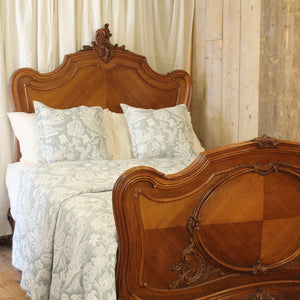 An ornate wooden bed frame with a carved headboard and matching footboard, dressed in pale blue and white patterned bedding against a rustic wood-paneled wall.
