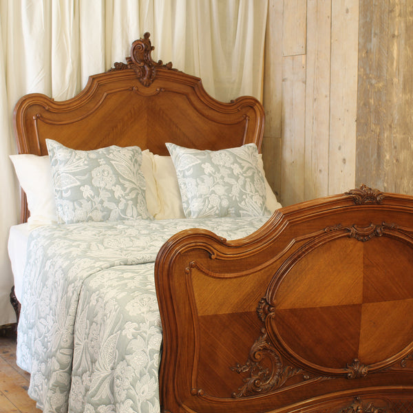 An ornate wooden bed frame with a carved headboard and matching footboard, dressed in pale blue and white patterned bedding against a rustic wood-paneled wall.