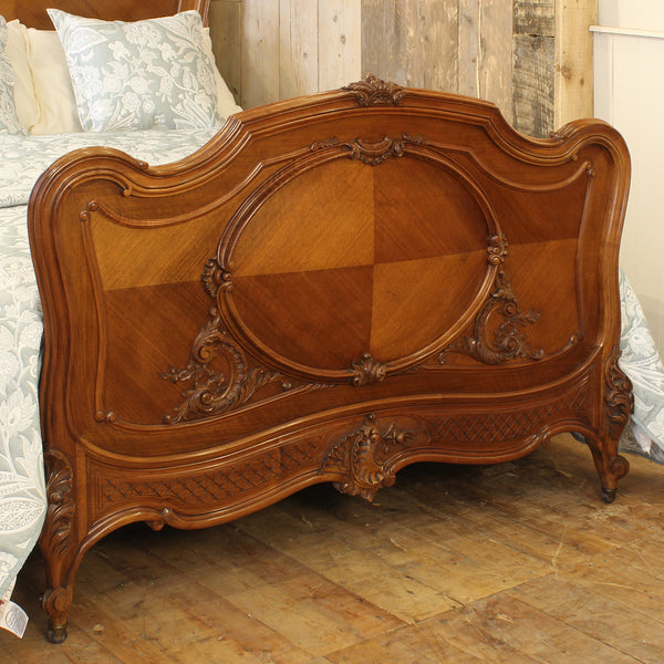An ornate, carved wooden bed headboard in a warm medium brown tone, positioned in a bedroom setting with a patterned white and blue quilt, rustic wooden wall, and wooden floor.