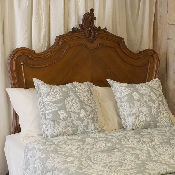 A close-up of a bed dressed in a light sage green and white floral damask patterned quilt and matching pillows, set against a carved dark wood headboard and cream curtains.