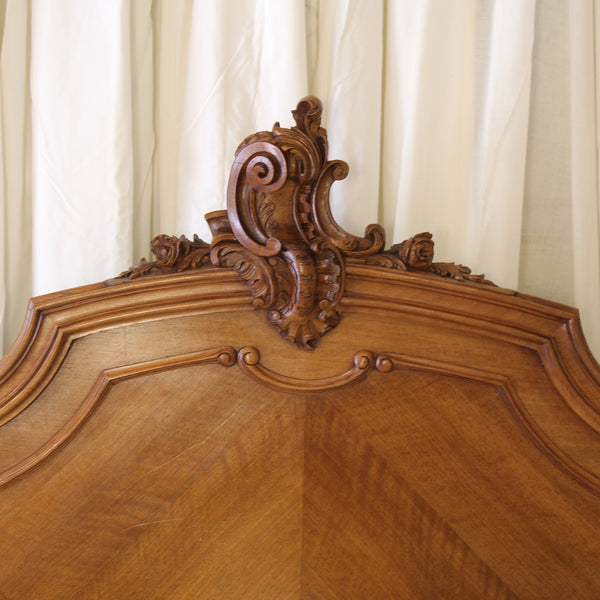 Close-up of an ornate wooden headboard with carved scrollwork and a curved panel, shown against cream curtains.