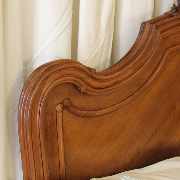Close-up of an ornate wooden headboard with carved scroll details, shown against draped white curtains and a white pillow.