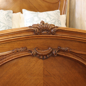 Close-up of an ornate wooden bed headboard with carved scrollwork and a warm medium-brown finish, with patterned pillows visible in the background.