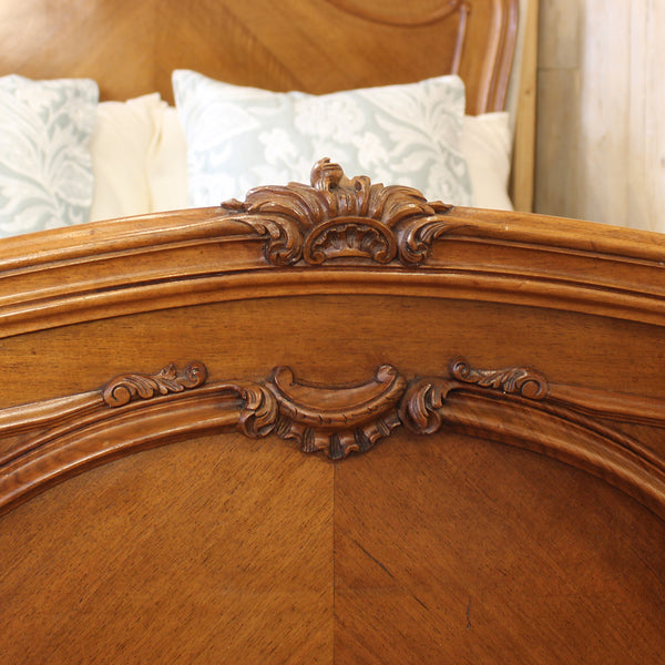 Close-up of an ornate wooden bed headboard with carved scrollwork and a warm medium-brown finish, with patterned pillows visible in the background.