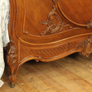 Close-up of an ornate carved wooden bed frame in warm medium brown wood, showing detailed scrollwork and a patterned panel, set against a rustic wooden floor with a white curtain visible on the left.
