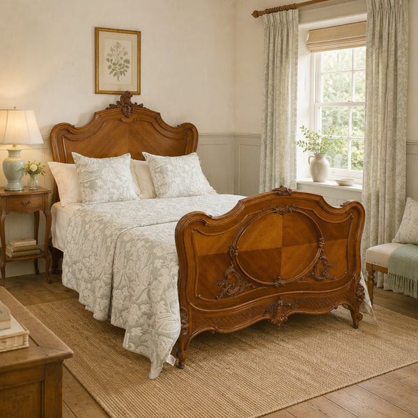 An ornate wooden bed frame with a carved headboard and matching footboard, dressed in light patterned bedding, shown in a softly lit bedroom with curtains, a window, and a side table.