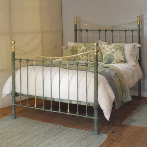 A vintage-style metal bed frame with a distressed sage green finish and polished gold-toned accents, shown fully made with white bedding and patterned green and blue pillows in a bedroom setting.