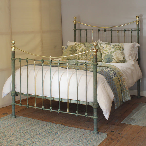 A vintage-style metal bed frame with a distressed sage green finish and polished gold-toned accents, shown fully made with white bedding and patterned green and blue pillows in a bedroom setting.