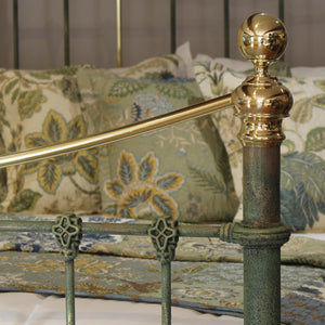 Close-up of an ornate metal bed frame with a distressed green-patinished post and polished brass rail and finial, shown in a bedroom setting with patterned bedding and pillows.