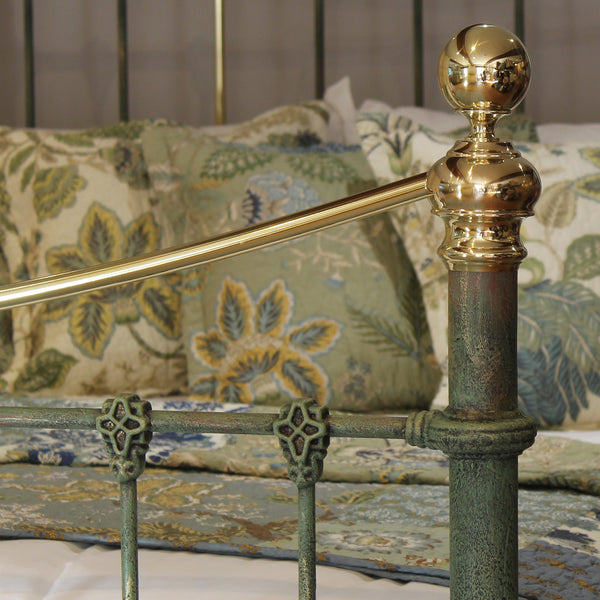 Close-up of an ornate metal bed frame with a distressed green-patinished post and polished brass rail and finial, shown in a bedroom setting with patterned bedding and pillows.