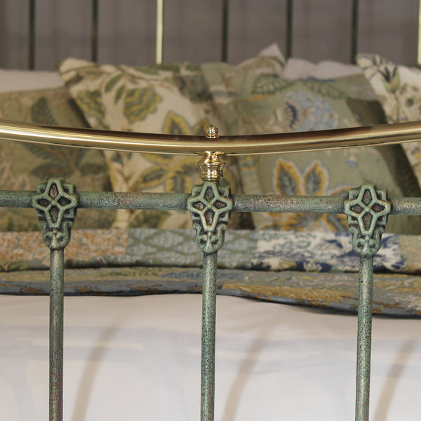Close-up of a metal bed frame with a distressed green/teal finish and a curved gold brass top rail, shown in a bedroom setting with patterned bedding in the background.