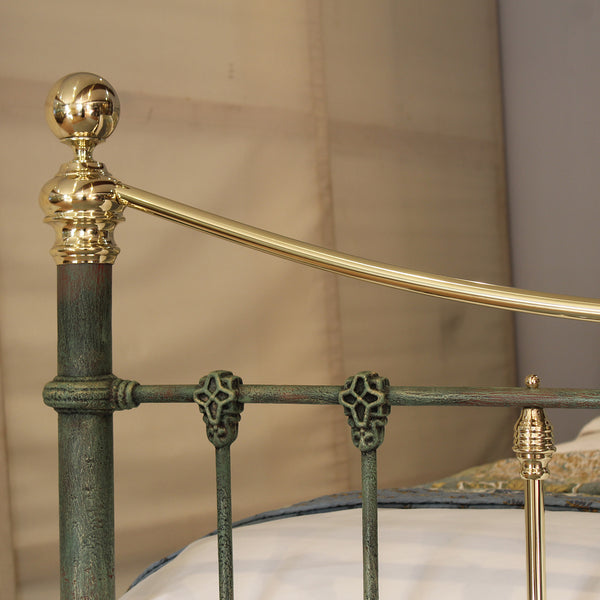 Close-up of an ornate metal bed frame with a distressed green-painted body and polished gold-toned accents, including a round finial on the post and decorative scroll brackets.