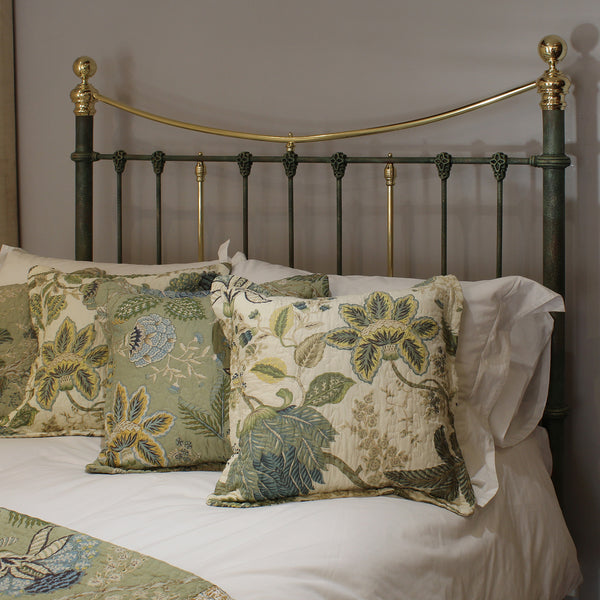 A close-up of a bed styled with white bedding and three decorative throw pillows featuring a botanical damask pattern in muted green, beige, and blue-gray tones, set against a dark green and gold metal headboard.