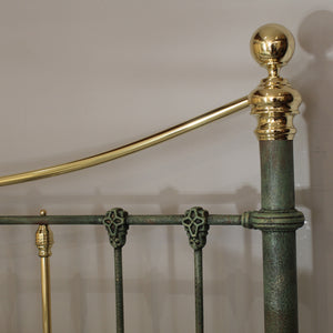 Close-up of an ornate metal bed frame showing a polished brass curved headboard bar, a round brass finial on the post, decorative skull-like bracket supports, and a distressed green-brass vertical post against a plain light background.