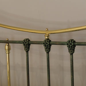Close-up of a metal bed frame showing dark green ornate vertical bars with decorative skull-like caps, a curved gold top rail, and matching gold finials against a plain light background.