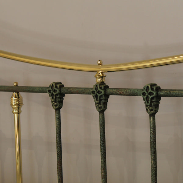 Close-up of a metal bed frame showing dark green ornate vertical bars with decorative skull-like caps, a curved gold top rail, and matching gold finials against a plain light background.