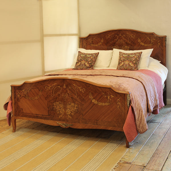An ornate wooden bed with intricate marquise inlay and floral carvings, dressed in pink and white bedding with two patterned accent pillows, set in a warm, neutral-toned bedroom.