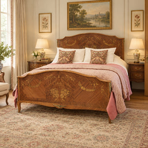 An ornate wooden bed frame with intricate marquetry and gold detailing, styled in a classic, symmetrical bedroom with matching nightstands, lamps, and a floral rug.
